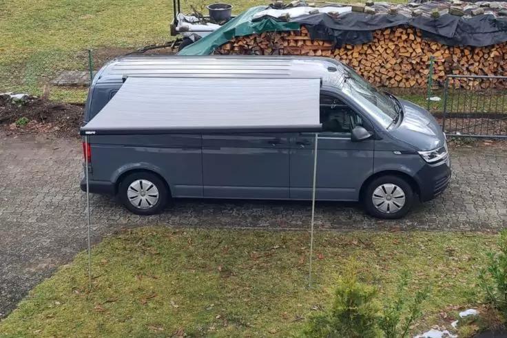 Seitansicht eines grauen Lieferwagens auf einem Schotterweg, Gras im Vordergrund, gestapeltes Brennholz mit Abdeckung im Hintergrund