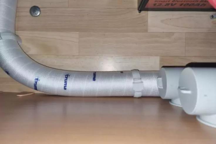 Silberner, isolierter flexibler Schlauch, verbunden mit wei&szlig;er Kunststoffverbindung, auf hellem Holzfu&szlig;boden vor einer Holzr&uuml;ckwand