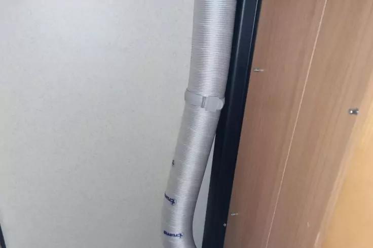 Vertikal stehendes hellgraues isoliertes Flexrohr, befestigt mit zwei Metallschellen, neben einer Holzschrankt&uuml;r an einer hellen Wand