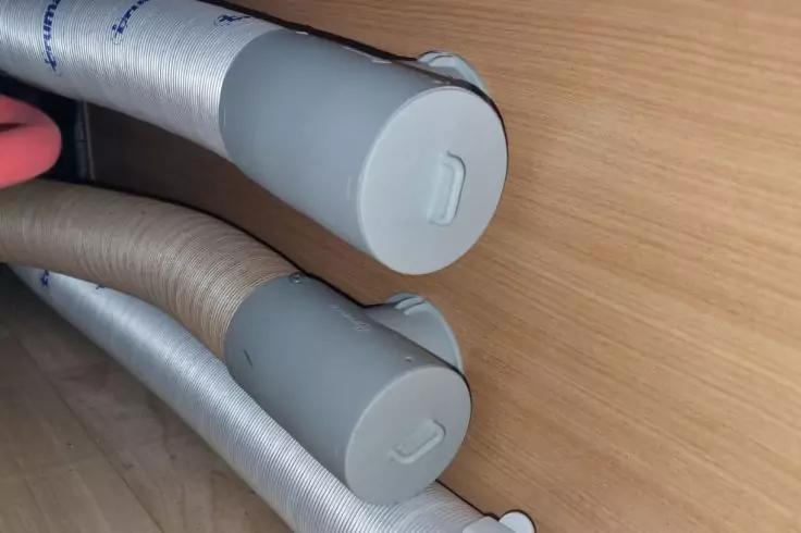 Zwei flexible Schl&auml;uche, einer silber, einer beige, mit grauen Endkappen liegen nebeneinander auf einem hellen Holzfu&szlig;boden vor einer Holzverkleidung