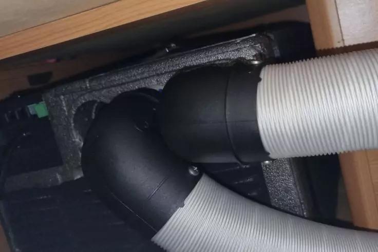 Zwei graue flexible Wellschl&auml;uche, verbunden mit einem schwarzen Kunststoffbogen, umgeben von grauem Schaumstoff, seitlich ein Holzrahmen sichtbar