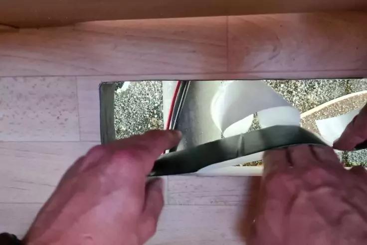 Zwei H&auml;nde halten einen schwarzen Streifen an eine rechteckige &Ouml;ffnung unter einer Holzverkleidung; darunter Schotterboden, ein wei&szlig;es Rohr und ein rundes wei&szlig;es Bauteil