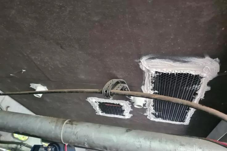 Dunkle Decke mit zwei rechteckigen schwarzen Gitter&ouml;ffnungen, umgeben von wei&szlig;em Dichtmaterial; ein gebogenes Kabel oder Rohr verl&auml;uft davor, darunter ein dicker Metallrohr