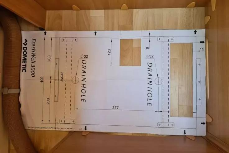 Innen eines hellen Holzgeh&auml;uses mit einer wei&szlig;en Papierschablone, aufgedruckt 'DRAIN HOLE', Ma&szlig;linien, Pfeile und zwei rechteckige Ausschnitte; links ein wei&szlig;er Schlauch; oben kleine Holzstifte