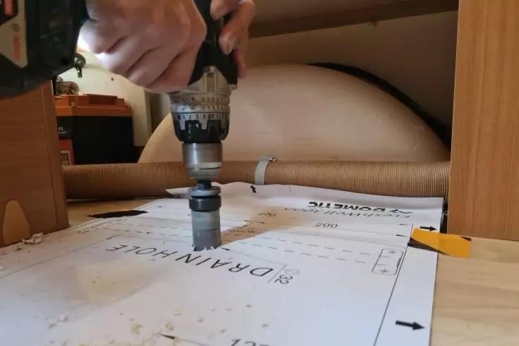 Hand h&auml;lt einen Akku-/Elektroschrauber mit Lochs&auml;geaufsatz, der in ein bedrucktes Papier auf dem Boden gedr&uuml;ckt wird; Holzunterseite eines M&ouml;belst&uuml;cks und ein rundes zylindrisches Polster/Rohr im Hintergrund