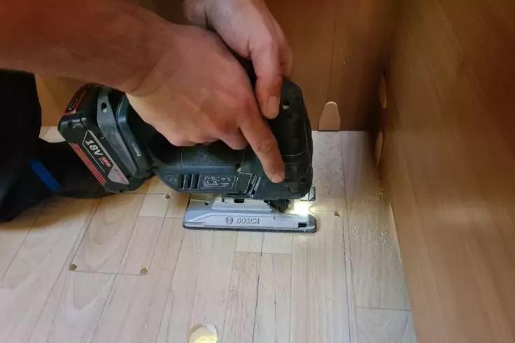 Zwei H&auml;nde halten eine Akku‑Stichs&auml;ge auf hellem Holzboden neben einer Holzverkleidung; ein rundes Loch im Boden und eine LED‑Leuchte an der S&auml;ge sichtbar