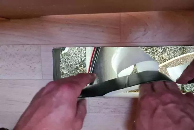 Zwei H&auml;nde halten eine schwarze Leiste an eine Metall-Schwelle zwischen Holzfu&szlig;boden und grauem Teppich; ein wei&szlig;es Kabel ist daneben sichtbar