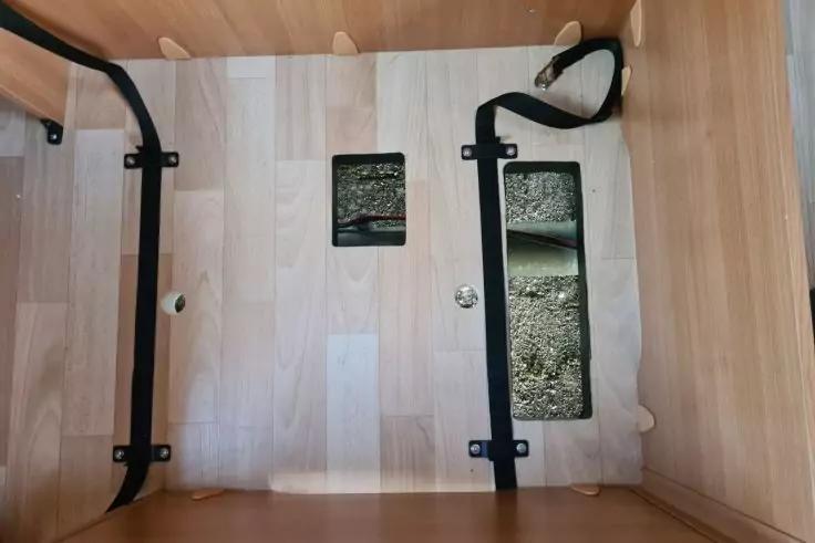 Innenraum aus hellem Holz mit zwei rechteckigen Aussparungen mit Kies, zwei schwarze Metallb&uuml;gel an den Seiten, schwarzer Gurt oben rechts, runde Metallverschraubung und kleines rundes Loch links