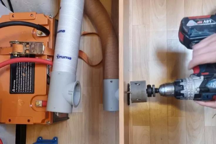 Hand h&auml;lt einen Akkuschrauber mit Lochs&auml;ge an einem Holzpaneel rechts; links ein orangefarbenes Ger&auml;t mit angeschlossenen Rohren und Schl&auml;uchen; Holzfu&szlig;boden sichtbar