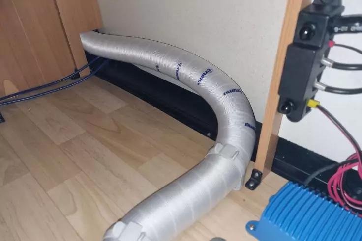 Silberner flexibler Schlauch auf hellem Laminatboden, entlang einer Wand gebogen; rechts Kabel und blaues Kunststoffger&auml;t, links ein Holzschrank