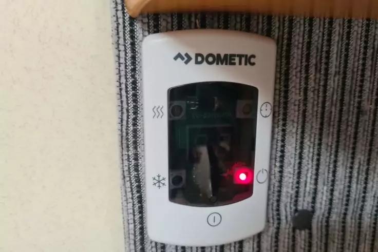 wei&szlig;es rechteckiges Ger&auml;t mit Schriftzug DOMETIC, dunklem Displayfenster und roter Leuchtdiode, montiert an beiger Wand neben gestreiftem Stoff, schwarzes Kabel unten rechts