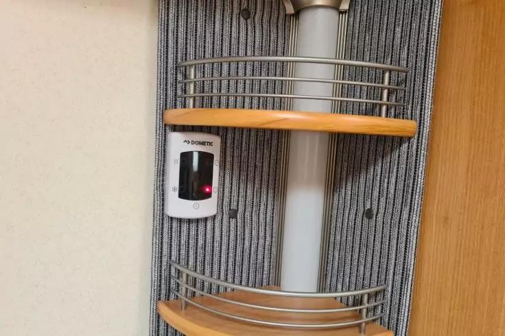 Drei h&ouml;lzerne Eckregalb&ouml;den mit Metallgel&auml;ndern an einem grauen, vertikal gerillten Paneel; mittig ein senkrechtes wei&szlig;es Rohr; links ein kleines wei&szlig;es Ger&auml;t mit roter Anzeige