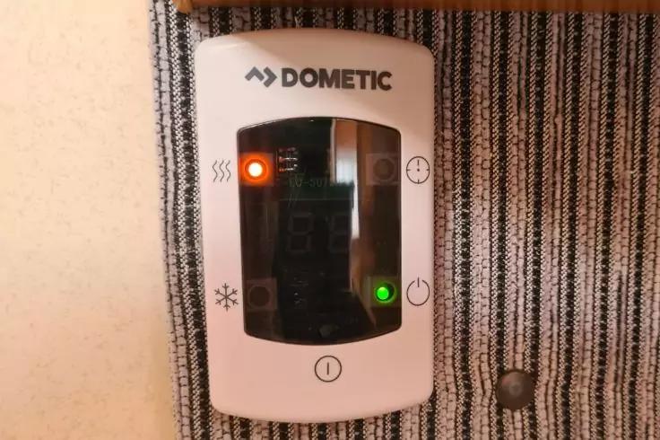 Wei&szlig;es Ger&auml;t mit DOMETIC-Schriftzug, schwarzem Displayfenster, orangeleuchtende LED links, gr&uuml;nleuchtende LED rechts, montiert auf grau-wei&szlig; gestreiftem Stoff