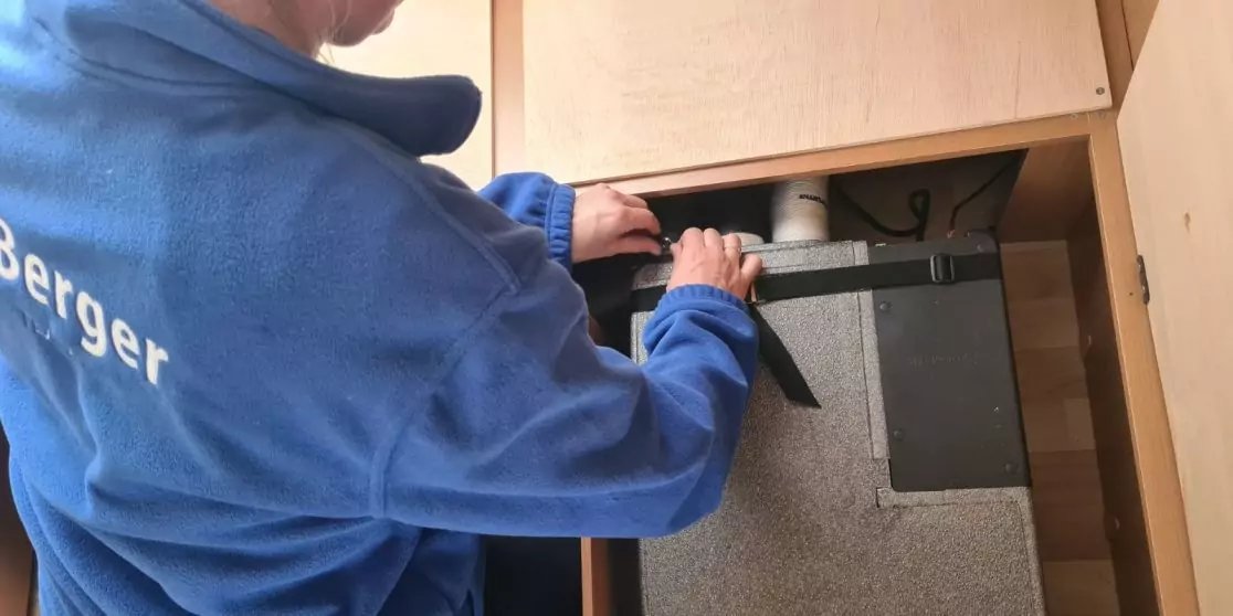 Person in blauem Pullover mit wei&szlig;em Schriftzug 'Berger', H&auml;nde an rundem L&uuml;ftungsrohr &uuml;ber einem rechteckigen Metallger&auml;t in einem Fach unter einem Holzschrank