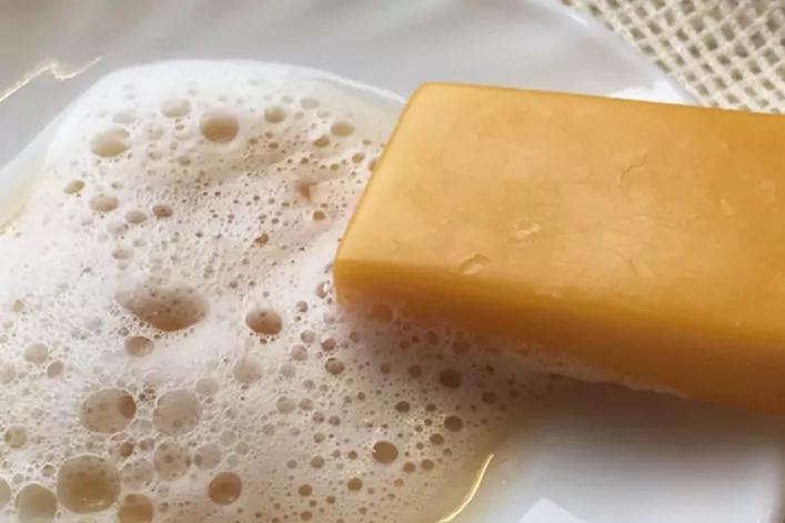 Gelbes Seifenst&uuml;ck auf wei&szlig;em Teller, Seifenschaum und gewebtes Tuch im Hintergrund