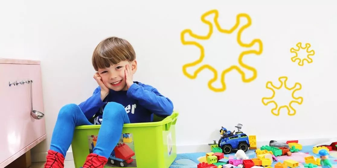 Ein Kind sitzt in einer gr&uuml;nen Plastikbox, H&auml;nde an den Wangen, l&auml;chelnd; rechts bunte Bausteine und ein Spielzeugauto, gelbe Wandaufkleber, links ein rosa Schrank