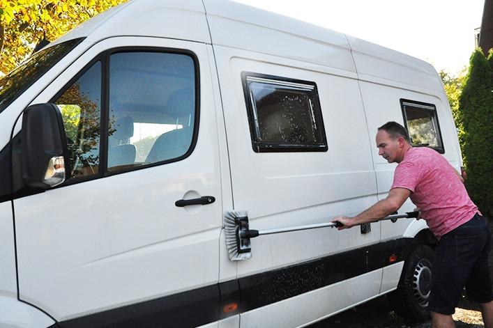 Mann in rosa T-Shirt zieht an einem horizontalen Metallgriff an der Seite eines wei&szlig;en Lieferwagens mit zwei rechteckigen Fenstern; B&uuml;sche und B&auml;ume im Hintergrund