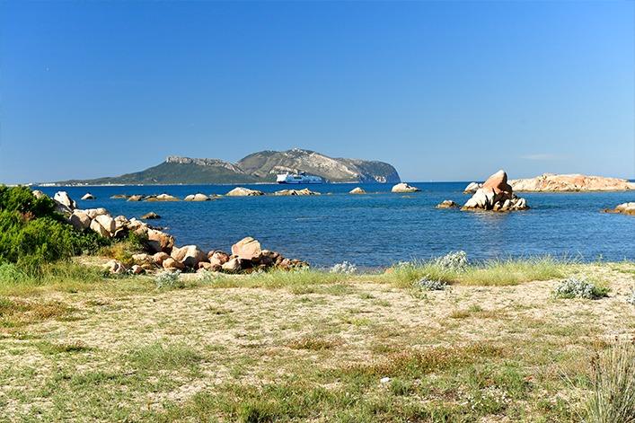 Gras- und sandboden im Vordergrund, felsige K&uuml;ste mit verstreuten Felsen im Meer, zwei felsige Inseln in der Ferne, blaues Meer und wolkenloser Himmel