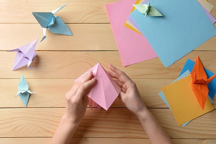 Zwei H&auml;nde falten ein rosafarbenes Origami; mehrere gefaltete Origami-V&ouml;gel und bunte Papierb&ouml;gen liegen auf einem Holztisch