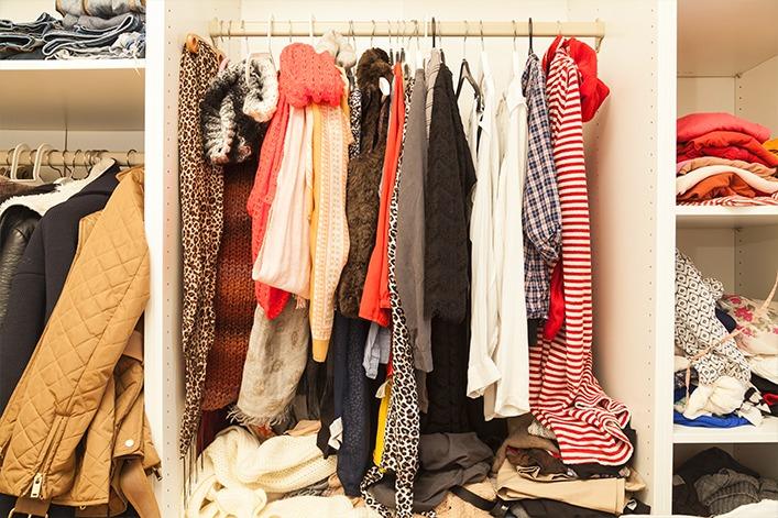 Offener Kleiderschrank mit an einer Stange h&auml;ngenden Jacken, M&auml;nteln, Schals, Hemden und Pullovern; gefaltete Kleidung und Stapel auf Regalen und Boden