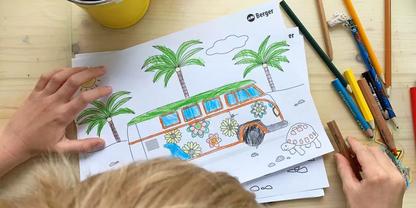 Zwei H&auml;nde und der obere Teil eines blonden Kopfes &uuml;ber einem Blatt mit Ausmalzeichnung eines dekorierten Kleinbusses, Palmen und einer kleinen Schildkr&ouml;te; Buntstifte und ein gelber Becher auf einem hellen Holztisch