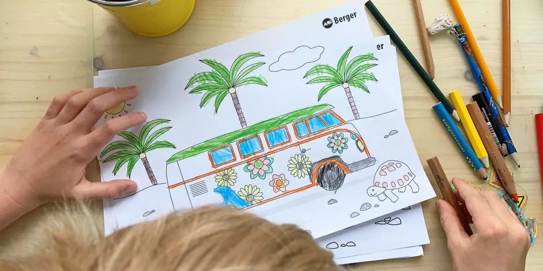 Zwei H&auml;nde und der obere Teil eines blonden Kopfes &uuml;ber einem Blatt mit Ausmalzeichnung eines dekorierten Kleinbusses, Palmen und einer kleinen Schildkr&ouml;te; Buntstifte und ein gelber Becher auf einem hellen Holztisch