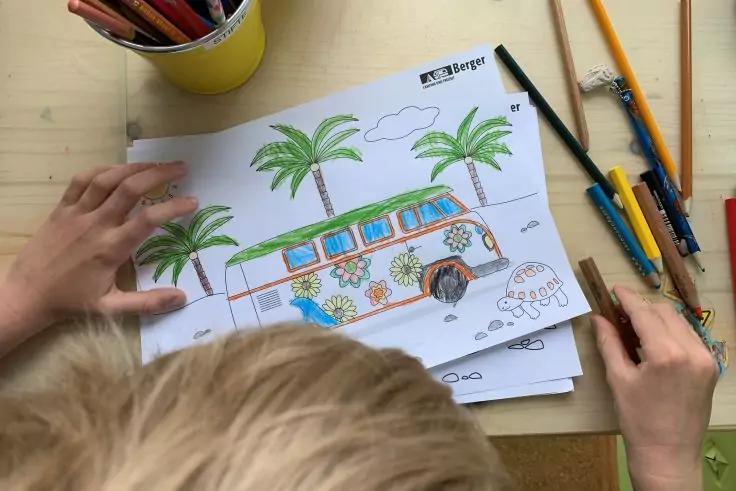 Person von hinten mit hellem Haar, H&auml;nde halten ein Ausmalbild eines bunt verzierten Busses mit Palmen; Buntstifte und ein Stiftebecher auf einem Holztisch