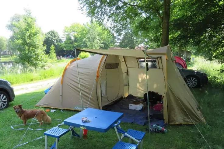 Campingzelt mit ge&ouml;ffnetem Vorzelt auf einer Wiese neben einem See, blaues Klapptisch-Set mit zwei Klappst&uuml;hlen, Hund liegt auf der Wiese, zwei Autos und B&auml;ume im Hintergrund