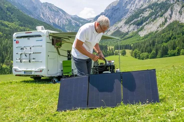 Eine Person richtet ein Solarpanel auf einer Wiese, im Hintergrund Wohnmobil, B&auml;ume und Berge