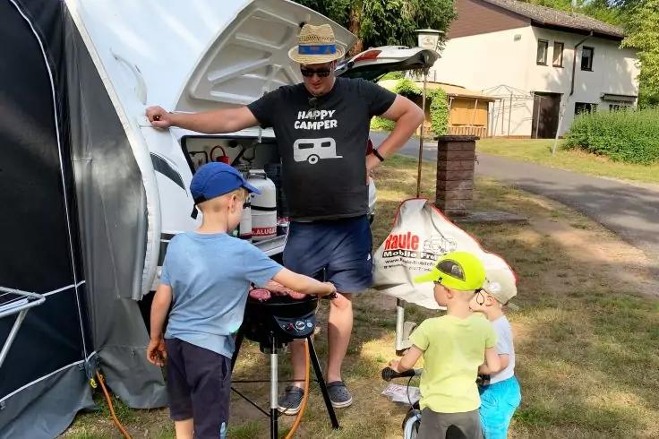 Mann mit Strohhut und schwarzem T‑Shirt mit Aufschrift HAPPY CAMPER steht vor einem ge&ouml;ffneten Wohnwagen; drei Kinder mit Kappen um einen kleinen Grill; H&auml;user und Rasen im Hintergrund