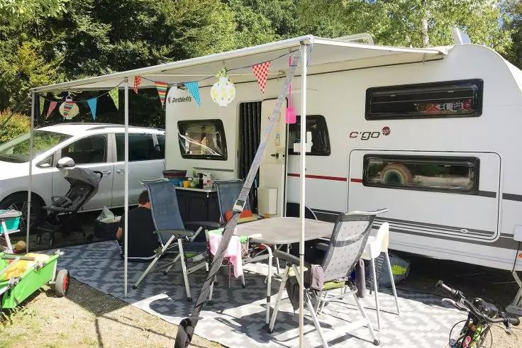 Wei&szlig;er Wohnwagen mit ausgefahrener Markise, Klapptisch und St&uuml;hle, Wimpelkette, geparktes Auto, Kinderfahrzeug und Fahrrad auf Rasenfl&auml;che vor B&auml;umen
