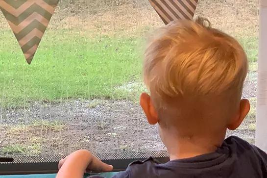 Kind mit kurzem blondem Haar von hinten, H&auml;nde auf Fensterbank, Blick auf Rasen und Kies drau&szlig;en, dreieckige Wimpel &uuml;ber dem Fenster