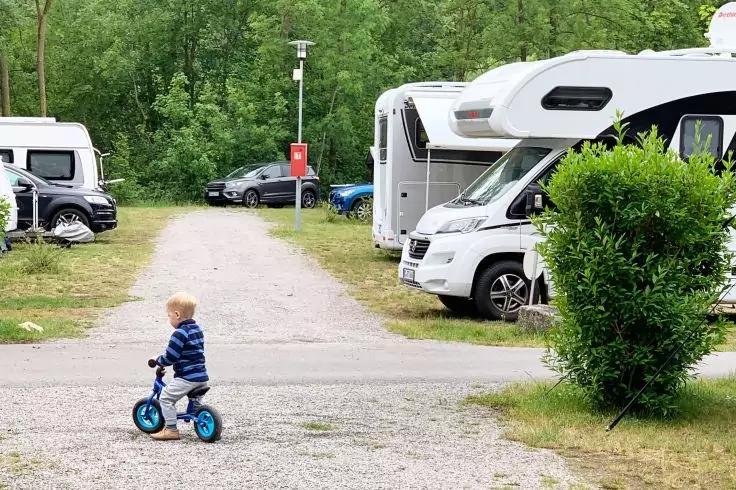 Kind auf blauem Laufrad auf Schotterweg zwischen geparkten Wohnmobilen und B&auml;umen
