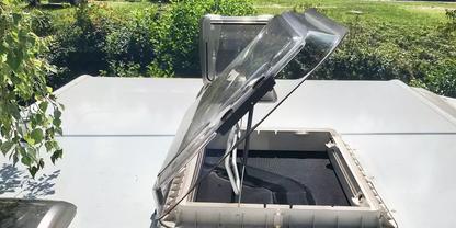 Aufgeklappte transparente Dachluke mit sichtbarem Scharnier und dunklem Innenraum auf einer flachen Dachfl&auml;che, im Hintergrund gr&uuml;nes Laub