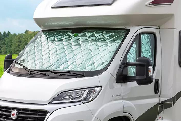 Vorderansicht eines weißen Wohnmobils mit gesteppter silberner Sonnenschutzfolie in der Windschutzscheibe; Außenspiegel, Scheinwerfer und Tür sichtbar; Bäume im Hintergrund