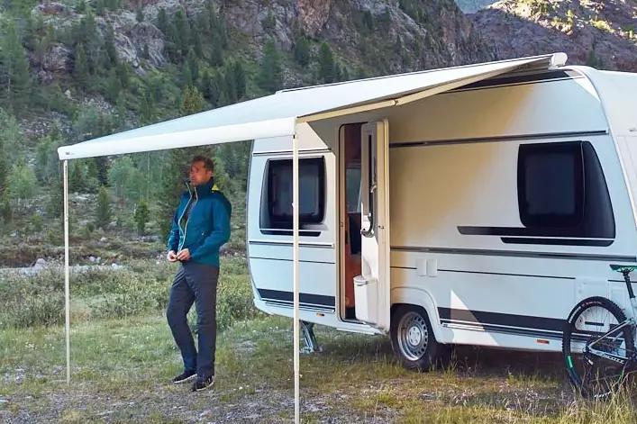 Person lehnt an einem Wohnwagen unter ausgefahrener Markise; geöffnete Tür, Fahrrad an der Seite, Grasfläche und bewaldete Berge im Hintergrund