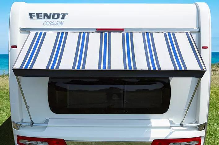 Weißer Wohnwagen mit blau-weiß gestreifter Markise über getöntem Heckfenster, Markenaufdruck FENDT, Gras und Meer im Hintergrund
