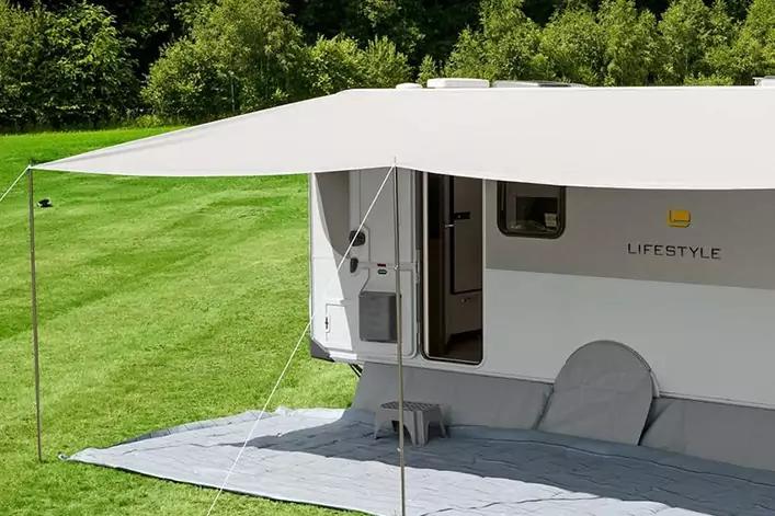Weißer Wohnwagen mit ausgefahrener Markise, offener Tür, Bodenmatte und kleinem Hocker auf einer Wiese vor Bäumen
