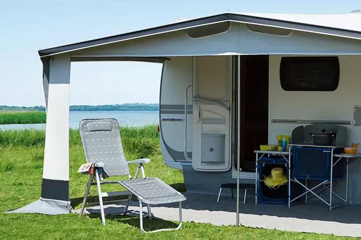 Wohnwagen mit ausgefahrener Markise auf Wiese am See, Liegestuhl, Campingtisch mit Stühlen und Ausrüstung