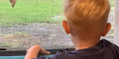 Person mit blonden Haaren von hinten, H&auml;nde auf Fensterbank, drau&szlig;en Gras und nasser Boden