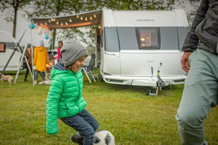 Kind in gr&uuml;ner Steppjacke und grauer M&uuml;tze tritt einen schwarz-wei&szlig;en Fu&szlig;ball auf einer Wiese vor einem wei&szlig;en Wohnwagen; Lichterkette am Vorzelt, weitere Personen im Hintergrund