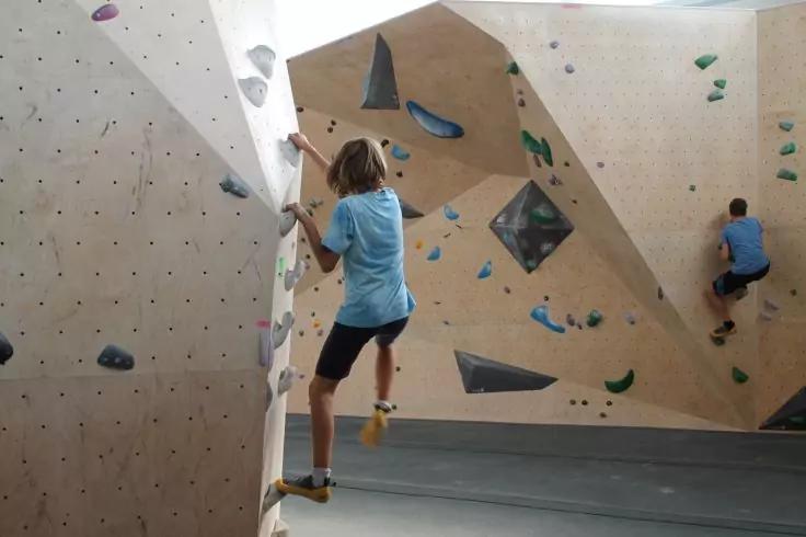 Zwei Personen in blauen Shirts bouldern an einer hellen Holz-Kletterwand mit bunten Griffen, graue Bodenmatten