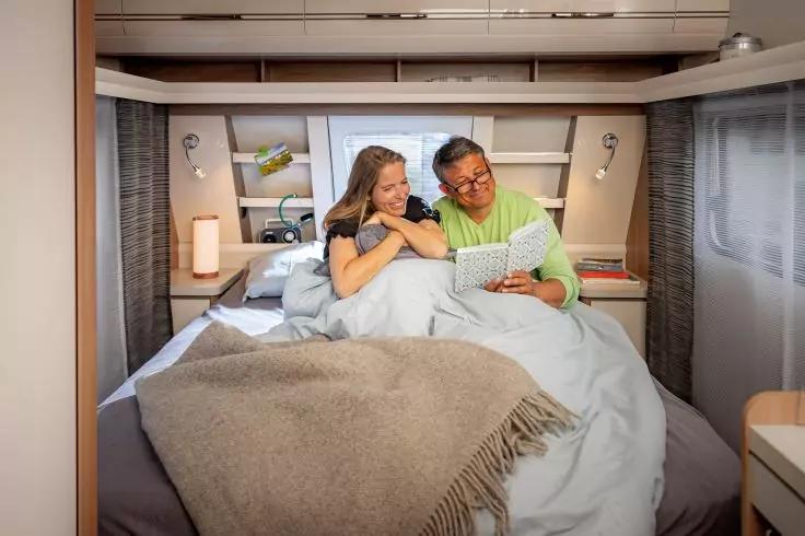 Zwei Personen sitzen in einem Bett im Inneren eines Wohnmobils, zugedeckt mit einer Decke und blicken auf ein Tablet; Regale und Leselampen sind im Hintergrund sichtbar