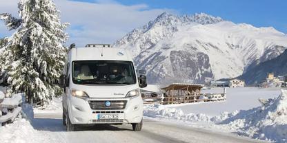 Wei&szlig;es Wohnmobil auf schneebedeckter Stra&szlig;e, Schnee an den Seiten, Tanne links, verschneite H&auml;user und Berge im Hintergrund, blauer Himmel