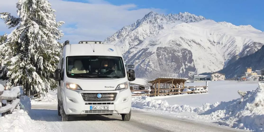 Wei&szlig;es Wohnmobil auf schneebedeckter Stra&szlig;e, Schnee an den Seiten, Tanne links, verschneite H&auml;user und Berge im Hintergrund, blauer Himmel