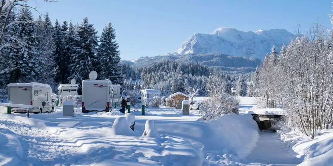 Mehrere Wohnmobile auf verschneiter Fl&auml;che, verschneite Wege und ein Holzbr&uuml;ckchen &uuml;ber einen Bach, Tannen und kleine verschneite H&uuml;tten, schneebedeckte Berge im Hintergrund unter blauem Himmel