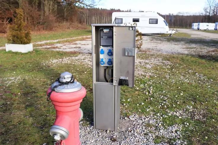 Roter Hydrant vorne links, daneben offener Metallschrank mit Steckdosen und Kabeln, im Hintergrund wei&szlig;er Wohnwagen, kiesiger Boden, Gras und Baumreihe