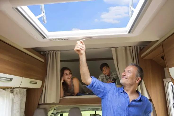 Wohnmobil-Innenraum: Mann reicht zur ge&ouml;ffneten Dachluke, Frau und Kind liegen im Alkoven, blauer Himmel sichtbar