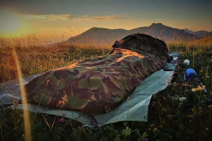 Camouflage-Schlafsack auf Isomatte in einer bl&uuml;henden Wiese, Berge im Hintergrund, Sonne niedrig am Horizont und zwei blaue Gegenst&auml;nde neben dem Schlafsack