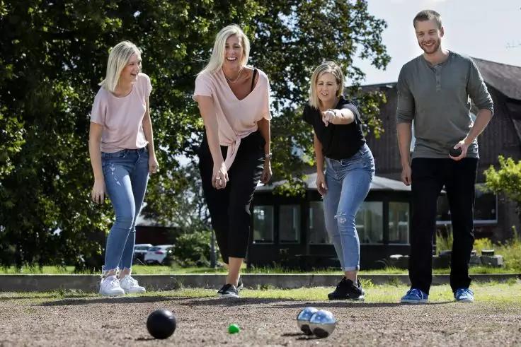 Vier Personen auf einem Schotterfeld; drei halten Kugeln; mehrere Kugeln liegen auf dem Boden; B&auml;ume und ein Geb&auml;ude im Hintergrund